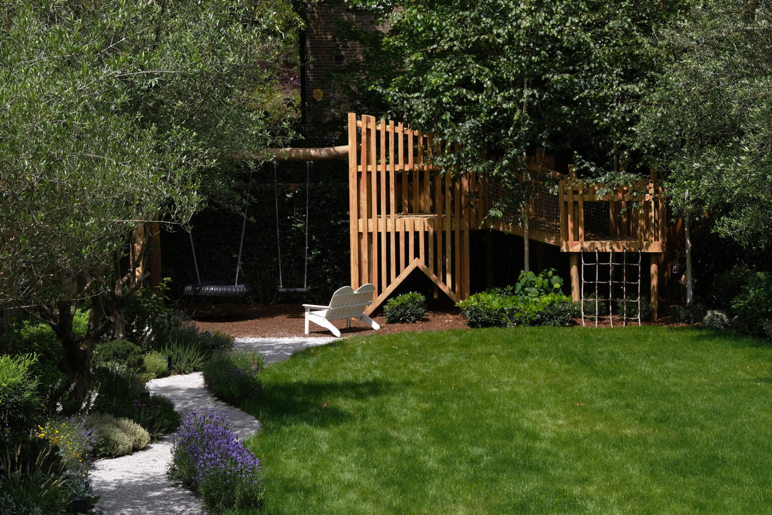 Family garden with a lawn, wooden play tower and shaded woodland planting