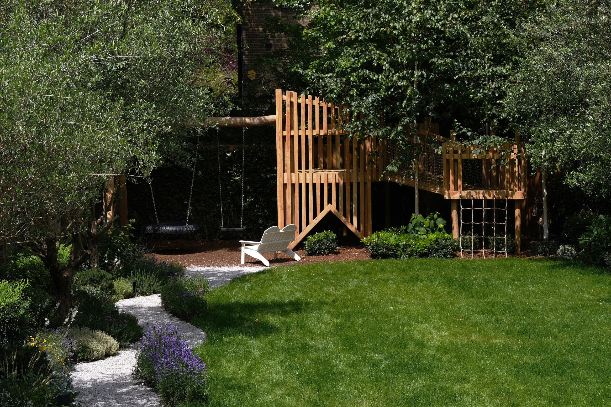 Family garden with a lawn, wooden play tower and shaded woodland planting